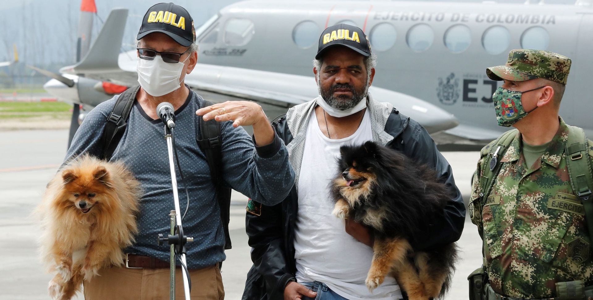 Zwei Männer mit kleinen Hunden auf dem Arm stehen auf einem Militärflugplatz vor einem Mikrophon und geben eine Pressekonferenz. Rechts steht ein kolumbianischer Soldat.