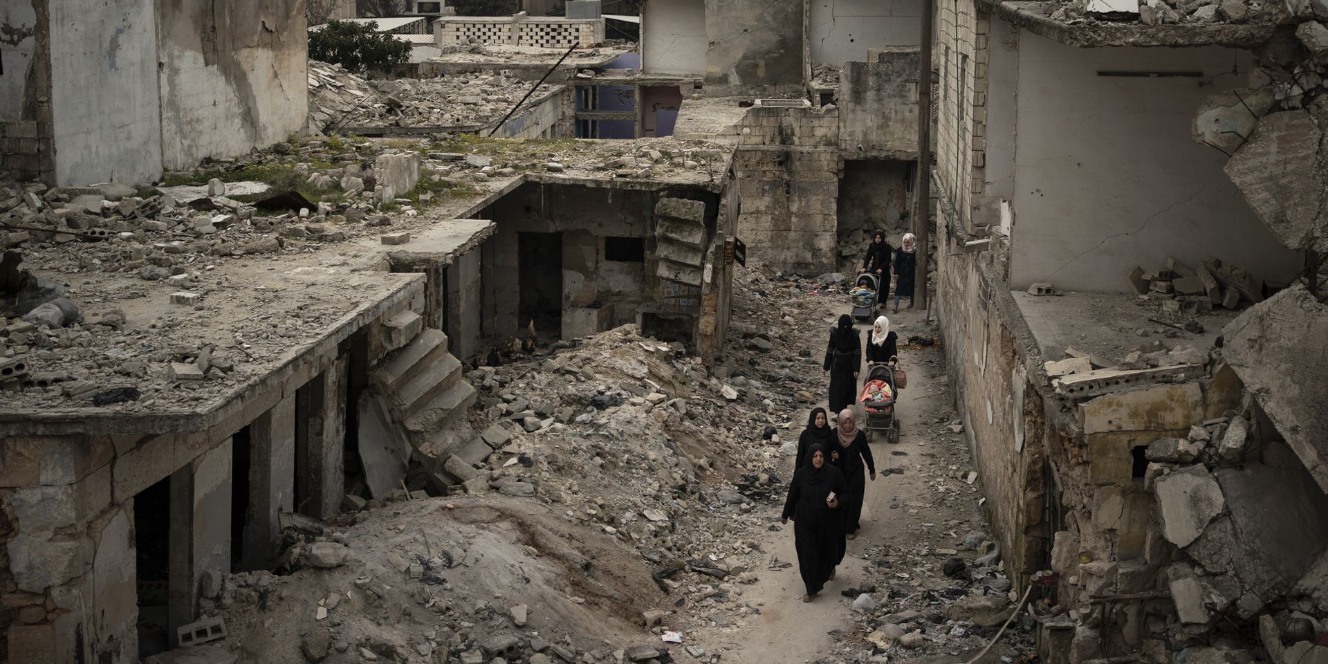 Women walk with prams through the rubble of a street with collapsed houses. 