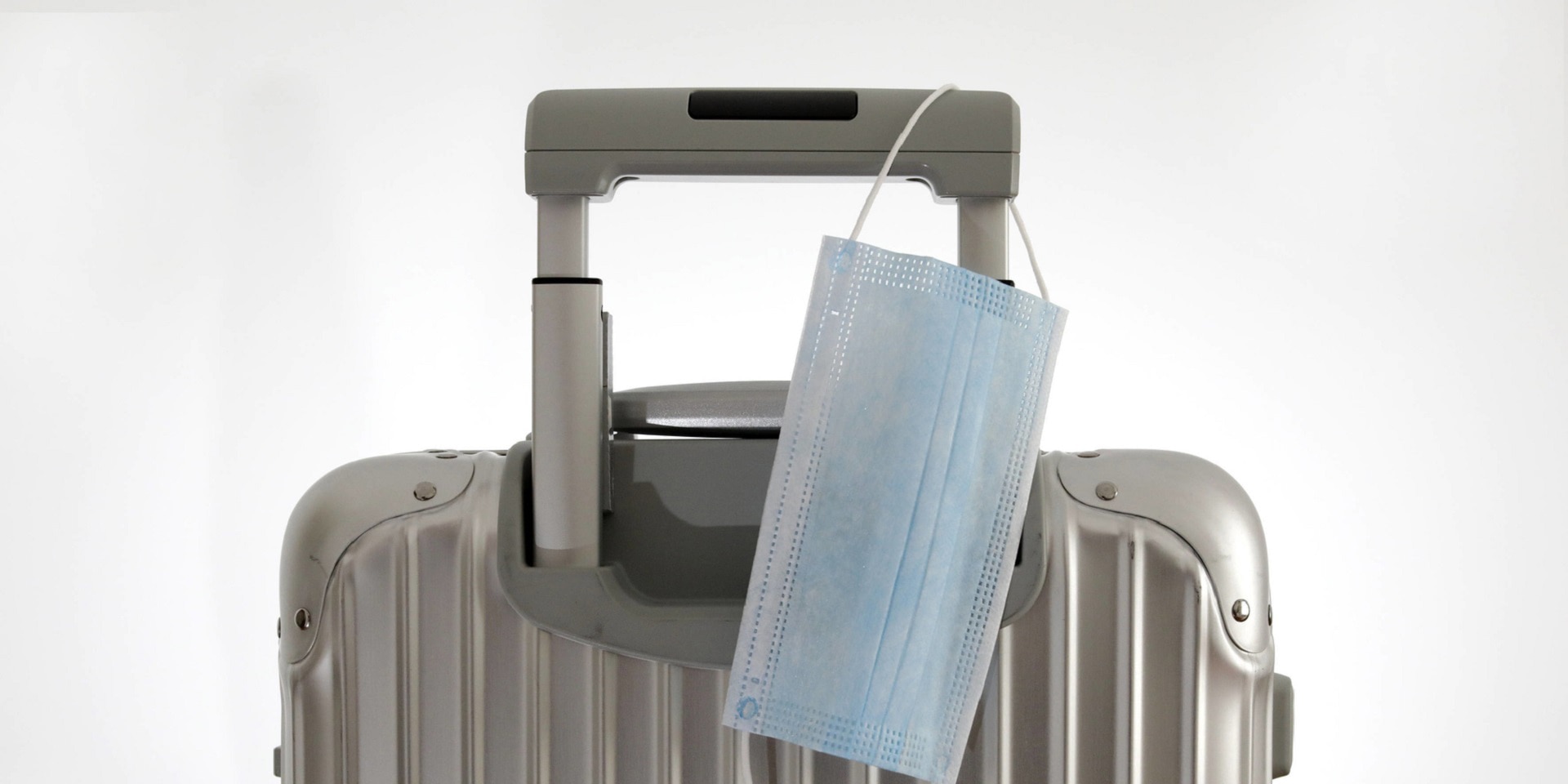A face mask hangs on the handle of a gray suitcase.
