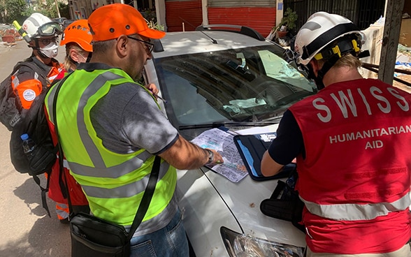 Four people stand by a damaged car and discuss a local map.