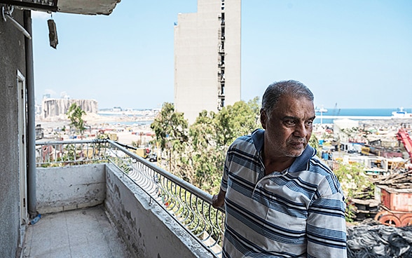 Ein Mann steht auf dem Balkon seines kaputten Wohnblocks und blickt in die Stadt. 