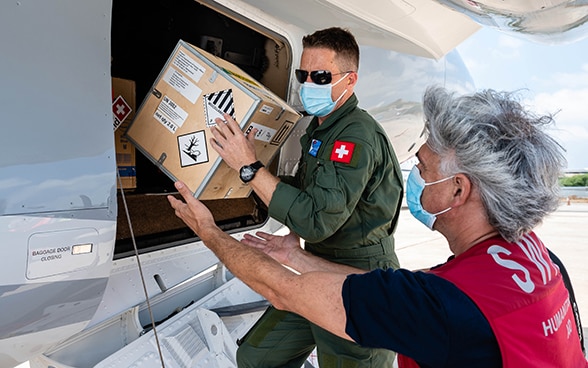 Blick auf einen Piloten der Luftwaffe, der gemeinsam mit einem Experten der Humanitären Hilfe des Bundes eine Kiste aus einem Flugzeug lädt.