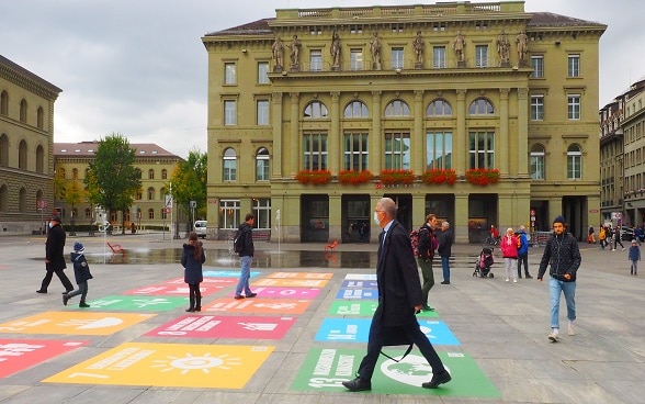 Passanten gehen über den Bundesplatz, auf dem Illustrationen der 17 Nachhaltigkeitsziele der Agenda 2030 zu sehen sind.