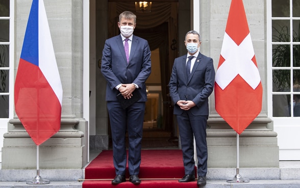 Ignazio Cassis and Tomáš Petříček are standing in front of an entrance, next to them the flags of their respective countries.