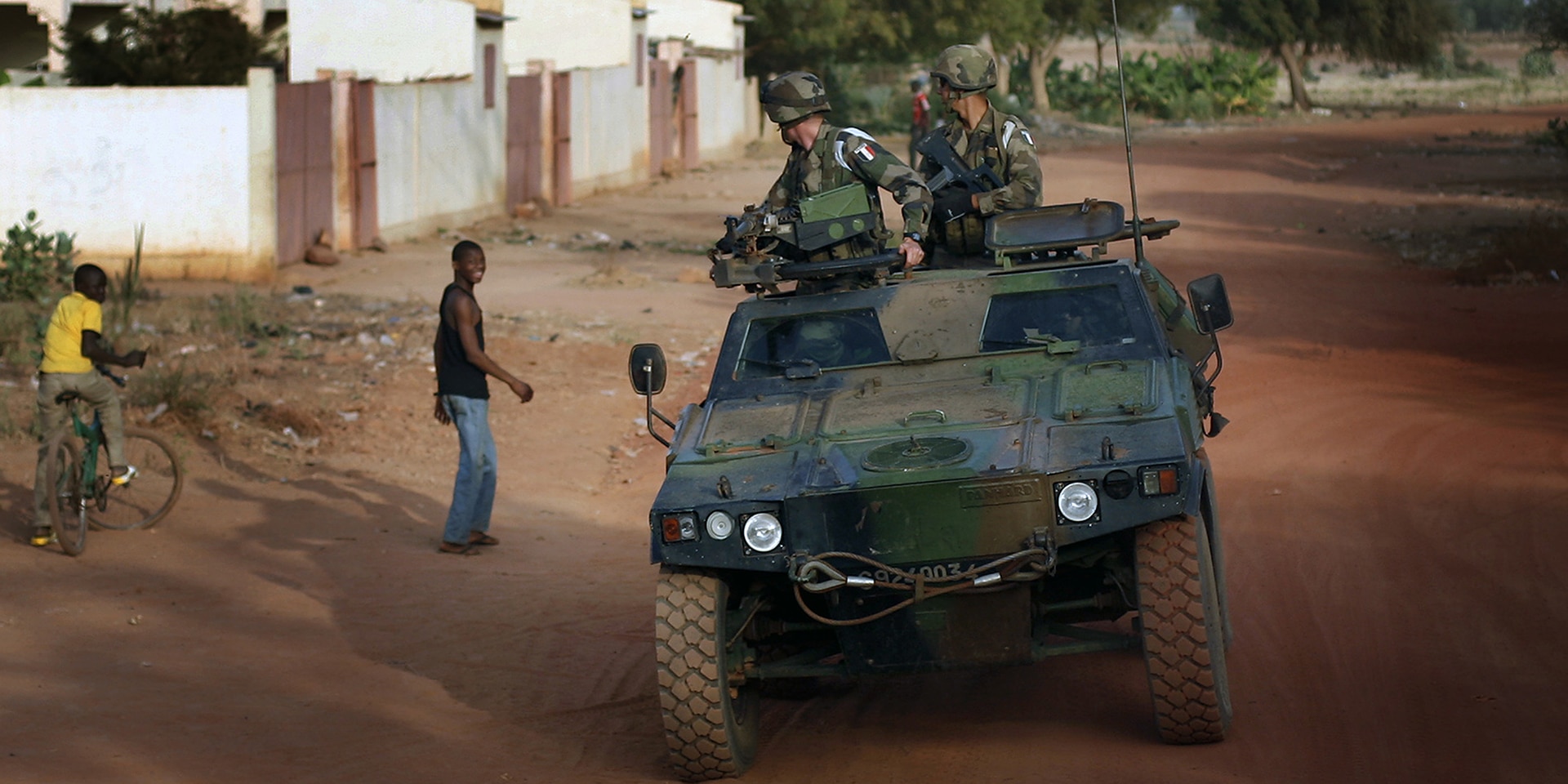 Un veicolo militare attraversa una strada di sabbia di un villaggio.