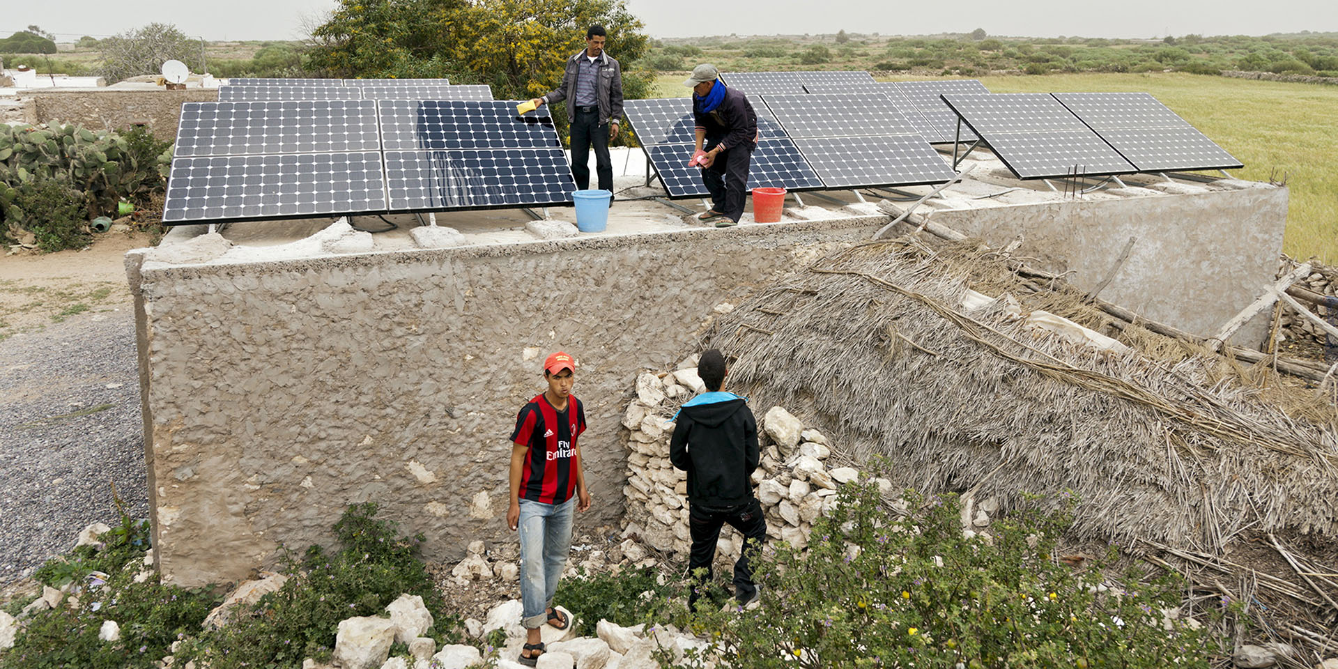 Two men stand on a stone building and wash solar panels. Next to the house are two other men.