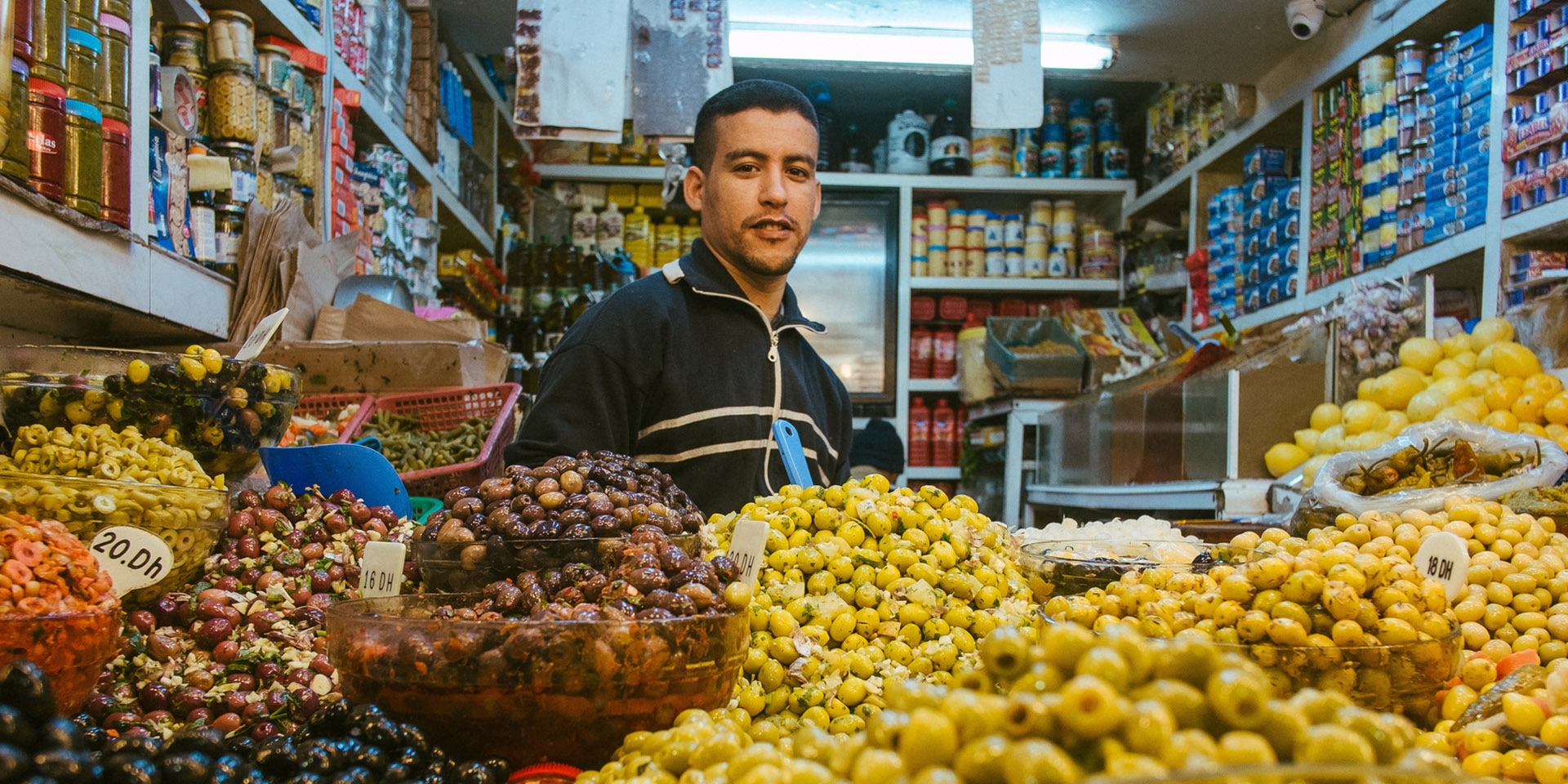 An olive seller stands in a small shop in front of a large display of olives of all kinds.