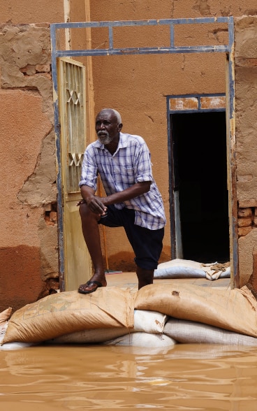 Un homme est debout sur des sacs devant sa maison à moitié détruite. La maison donne sur la route submergée par les eaux.