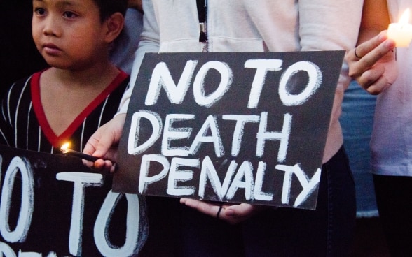 In the foreground, a child stands next to an adult who is holding up a sign saying "No to death penalty".