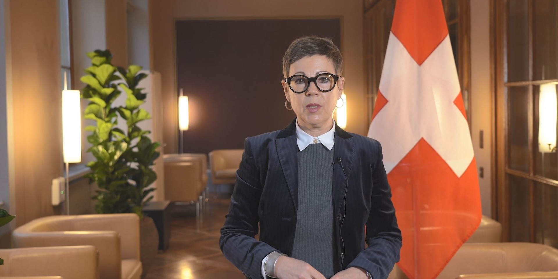 FDFA State Secretary Krystyna Marty Lang stands in a wood-panelled room and speaks into a camera. The Swiss flag can be seen in the background.