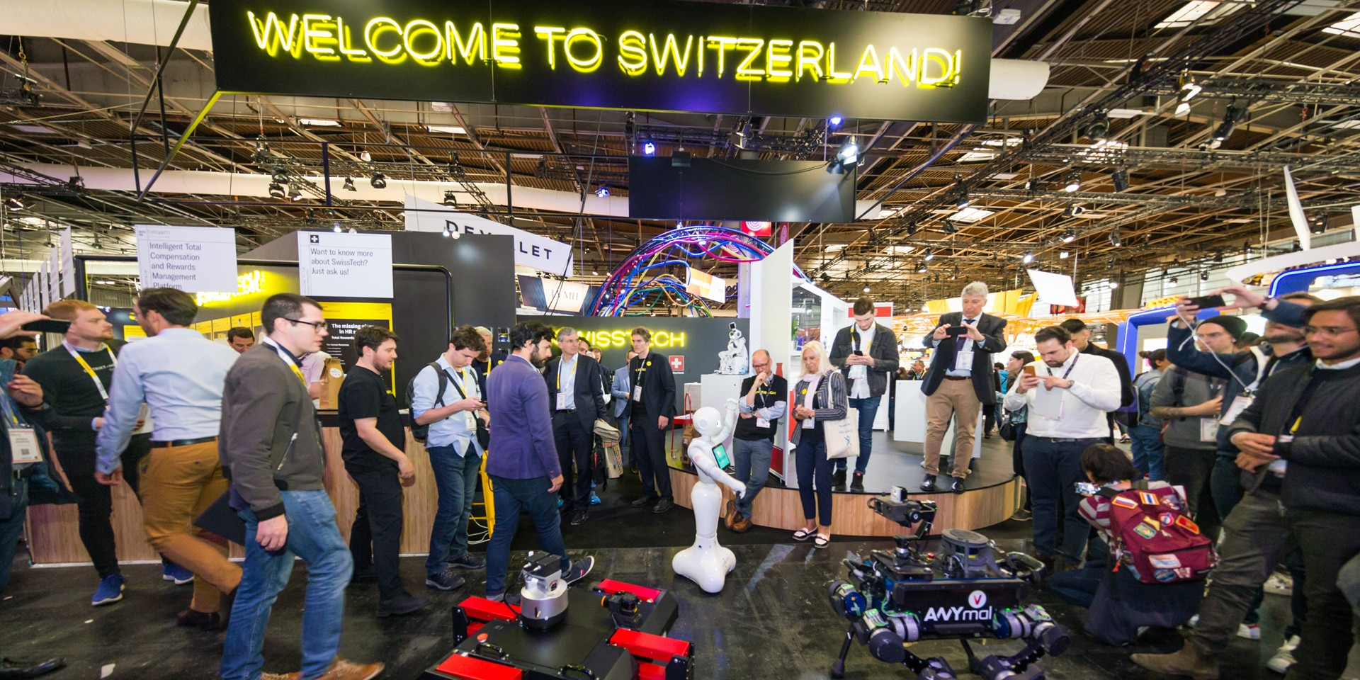 In an exhibition hall, people look at drones, robots and innovative technologies. Above them is a sign reading "Welcome to Switzerland".