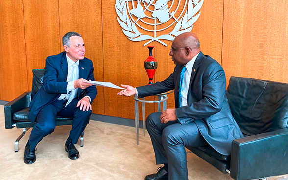 Against the backdrop of the UN emblem, Federal Councillor Ignazio Cassis hands over a document to the President of the UN General Assembly Abdulla Shahid (both seated).