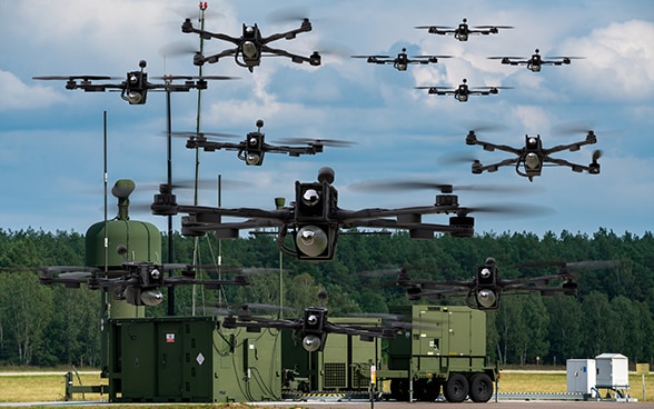 Zahlreiche Drohnen fliegen über militärischen Einrichtungen vor einem Wald. 