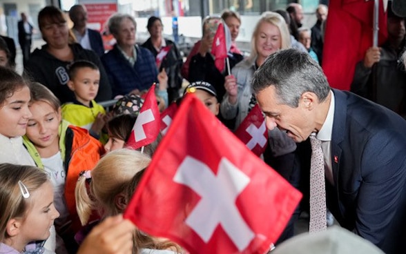 Il binario della stazione ferroviaria di Airolo è gremito di persone che accolgono il treno speciale. Il presidente della Confederazione Ignazio Cassis saluta i presenti mentre gli alunni della scuola sventolano le bandierine svizzere.  