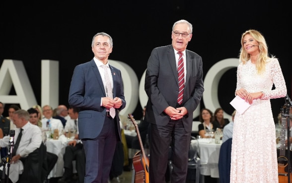 Ignazio Cassis, Guy Parmelin and Christa Rigozzi speak on a stage.