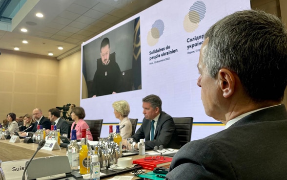 Bundesrat Cassis sitzt am Tisch und hört dem ukrainischen Präsidenten Selenski zu, der per Video auf einer Leinwand zu sehen ist.