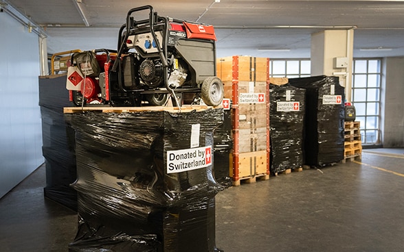  In a warehouse, a box with a picture of a mobile heater on a pallet is in the foreground. In the background are more pallets on which packed electricity generators are placed.