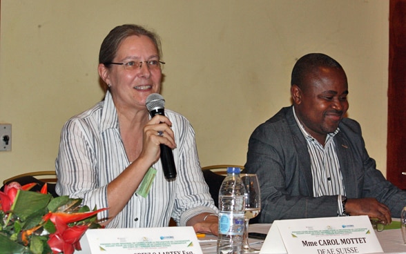 Carol Mottet speaking into a microphone at an event, sitting next to an African man.