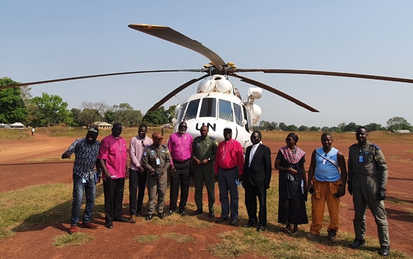 Ten African men and one woman stand in front of a white UN helicopter on sandy ground.