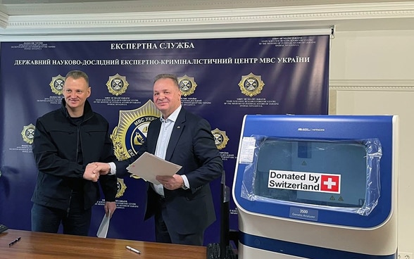 Ambassador Claude Wild shakes hands with a representative of the Ukrainian authorities. In the foreground are DNA analysis machines with the sticker "Donated by Switzerland".