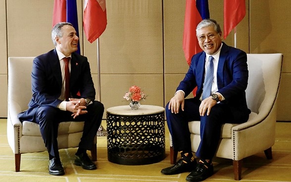 Ignazio Cassis et Enrique Manalo sont assis sur deux chaises. Derrière eux, les drapeaux de leur pays respectif.