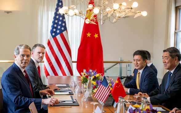 Die Delegationen der USA und Chinas sitzen an einem hölzernen Tisch. Im Hintergrund sind die Flaggen der beiden Länder zu sehen. 