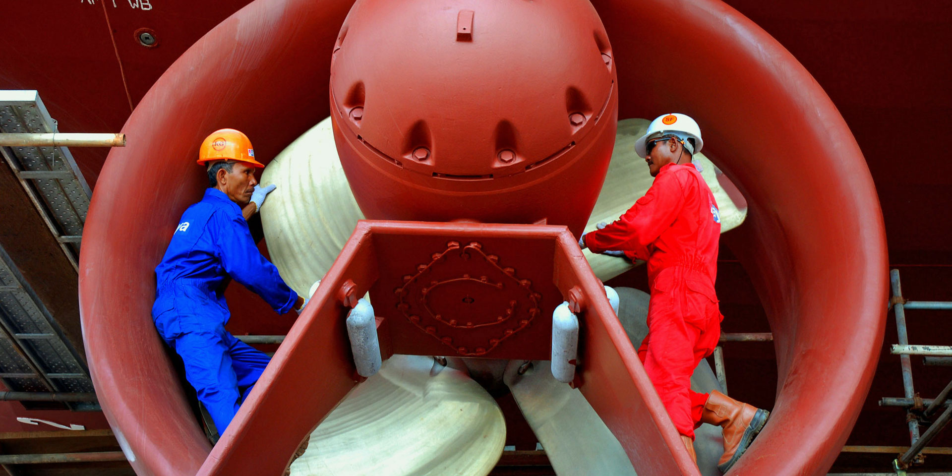 Two workers in blue and red overalls are working on a reactor in a hangar.
