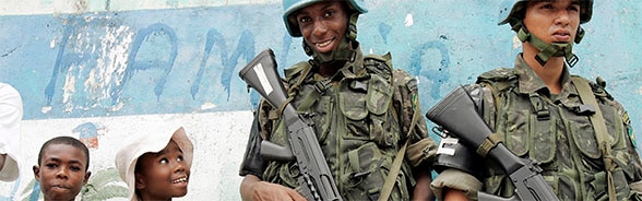 Zwei UNO-Soldaten mit umgehängter Waffe neben lächelnden Kindern an einer Veranstaltung für die lokale Bevölkerung in Haiti