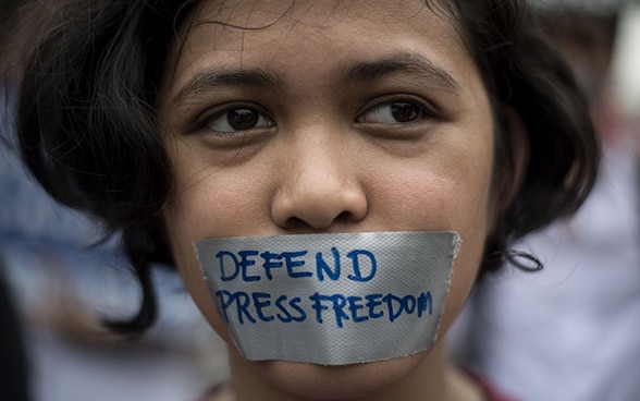 Portrait d’une petite fille dont la bouche est recouverte d’un morceau de ruban adhésif sur lequel sont inscrits les mots «defend press freedom» (défendons la liberté de la presse).