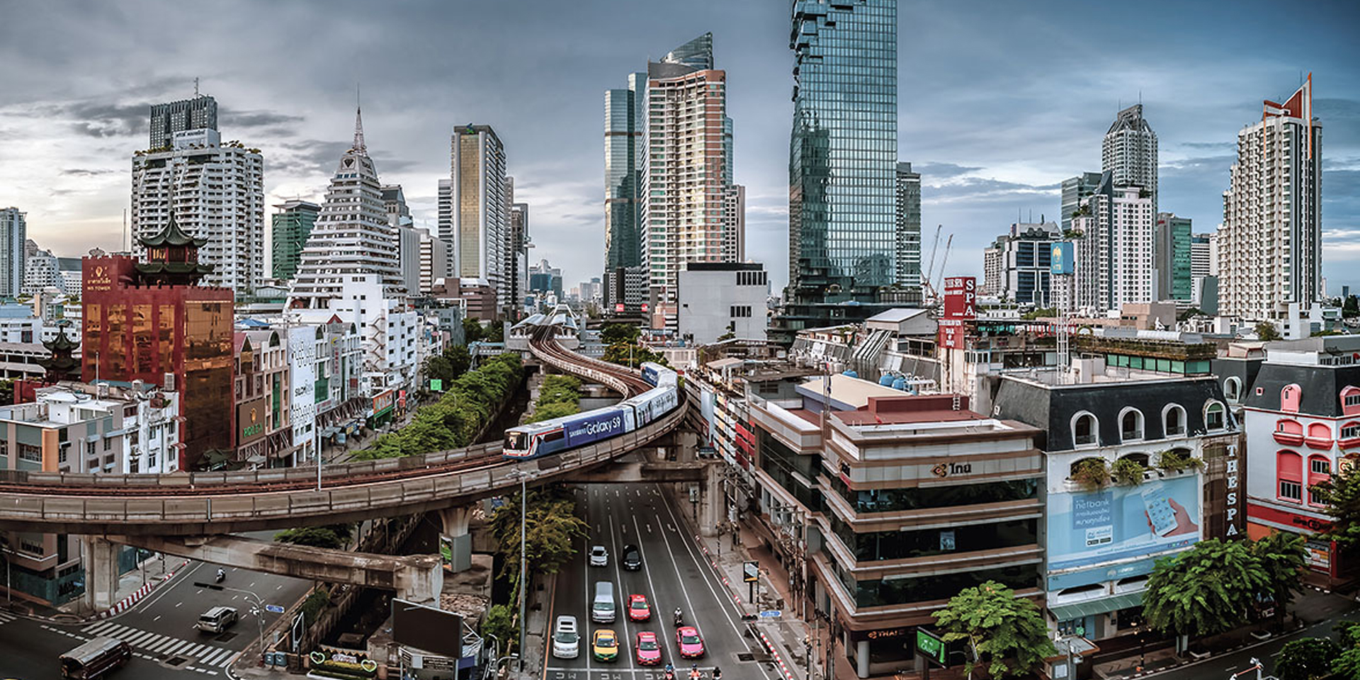  An aerial metro passes over a road with skyscrapers in the background.