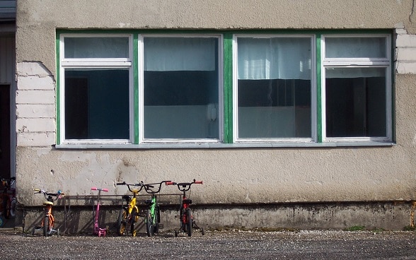 Vier Kinderfahrräder stehen vor einem Schulhaus. Die graue Fassade bröckelt, die Fenster sind einfach verglast.