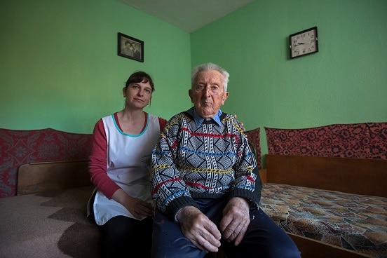 Un anziano è seduto su un sofà di casa sua assieme alla persona che lo assiste.
