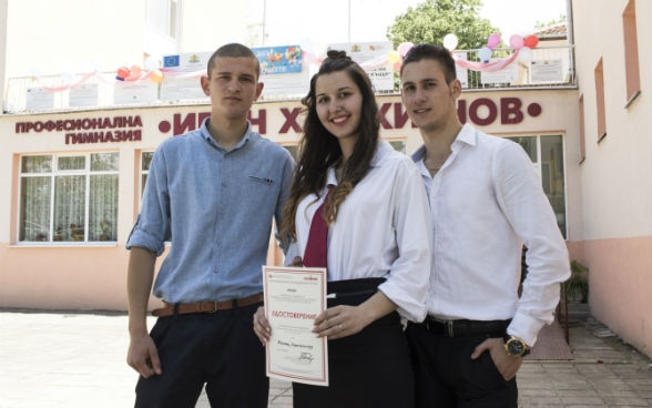 Three students, two boys and a girl, who holds a certificate, stand in front of their school.